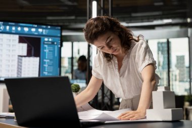Kadın emlak mühendisi ofisteki dizüstü bilgisayarın detaylarını araştırıyor, kentsel yenilik merkezindeki bir inşaat projesi için kentsel gelişim üzerinde çalışıyor. İnşaat alanı.