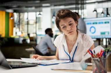 Ciddi bir kadın muhasebeci ofis yakınlarında dizüstü bilgisayarla finansal belgeleri okuyor. Masasında kağıtları olan beyaz bir kadın şirket değerlendirme raporlarını hazırlıyor..