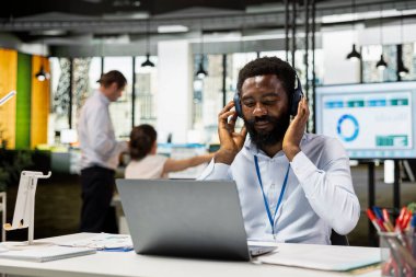 Bilgisayarı kullanan ve müzik dinleyen bir ofis çalışanı, internet üzerinden şirket güncellemelerini kontrol ediyor. İşadamı kulaklıklara dokunuyor, müzik listesinin keyfini çıkarırken aynı zamanda kurumsal çalışma alanındaki bütçe raporlarını tamamlıyor.