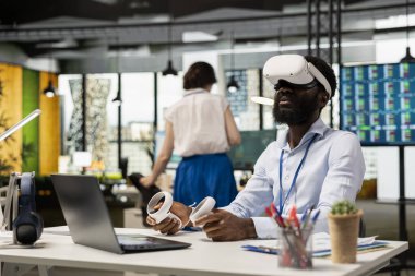 Siyahi erkek çalışanlar proje brifingi ve pazarlama stratejileri için AR kulaklığı kullanıyor. VR gözlüklü Afrikalı Amerikalı işadamı laptopta çalışıyor, iş yerindeki belgeleri inceliyor..