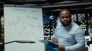 Erkek danışman, raporları incelemek, aylık verileri toplamak ve risk değerlendirmesi ve araştırma için projeler hazırlamak üzere beyaz tahtaya şamdan çizimleri çiziyor. Öngörü.