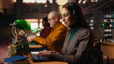 Yetişkin bir öğrenci kütüphanede dizüstü bilgisayar dinleyip notlar alıyor ve online ders sırasında araştırma yapıyor. Diplomasını almak için bir uzmanla uzaktan eğitim görüyor..