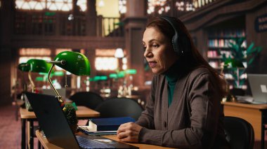 Yaşlı kadın üniversite öğrencisi Webinar 'da profesörle bilgisayar başında iletişim kuruyor, klasik akademik araştırmalarla modern telekomünikasyon kütüphanesinde okuyor. Seminer dersi..