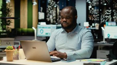 Siyahi işadamı saati kontrol ediyor ve ofisteki masa lambasını kapatıyor, yeni bir bakış açısı için strateji kuruyor ve işlerini bitiriyor. Mesai saatleri dışında mesai yapan biri. Kamera B.