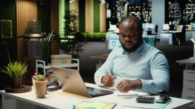 Bir sunum için el yazısı notları hazırlayan siyah işletme uzmanı, finans belgelerinden gerekli verileri çıkartıyor ve iş hedefleri ve görevleri için güncellemelerle özet geçiyor. Kamera A.