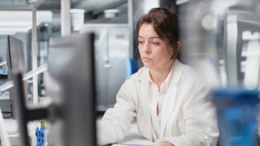 İş yerindeki Asyalı iş arkadaşının yanındaki laboratuvar camlarını gösterge çözeltisiyle dolduran kimyager. Laboratuar bilimcisi, bilgisayar ve kamera B kullanarak, meslektaşının yanında kimyasal sıvı bulunan silindiri inceliyor.