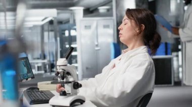 Yorgun laboratuvar bilim adamı gözlerini ovuşturuyor, PC kullanarak genetik işaretleri değerlendirmekten yoruldu. Yorgun uzman, bilgisayar masasındaki DNA iplikçikleri, iç çekmeler, kamera B...