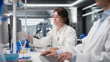 İş yerindeki Asyalı iş arkadaşının yanındaki laboratuvar camlarını gösterge çözeltisiyle dolduran kimyager. Laboratuar bilimcisi, bilgisayar ve kamera A 'yı kullanarak meslektaşının yanında kimyasal sıvı bulunan silindiri inceliyor.