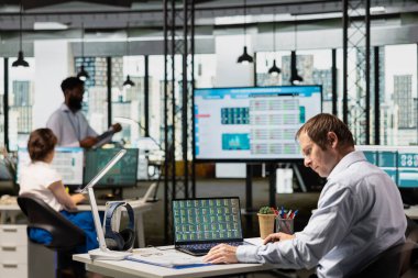 Ofisteki erkek analist, panodaki bilgilerle karşılaştırarak dijital cihazdaki hisse alım verilerini inceliyor. İşadamı önx piyasasını analiz etmek ve belgelerdeki indeks çizelgelerini incelemek için dizüstü bilgisayar kullanıyor.