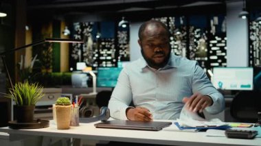 Siyahi genç işadamı gece yarısı görevlerini tamamlıyor ve lambasını kapatıyor. Kompleks proje için fazla mesai yaptıktan sonra ofisinden aşırı yüklenmiş ve yorgun bir şekilde ayrılıyor. Kilitliyorum. Kamera A.