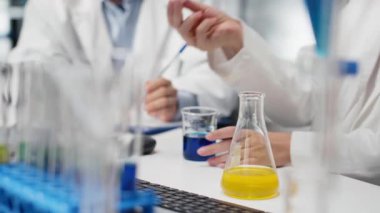 Laboratuar müdürü ve stajyer, araştırma merkezindeki yeşil gösterge çözeltisiyle dolu cam bardakları sallıyor. Silindir içinde kimyasal sıvıları karıştıran çok ırklı bir ekip, kamera B