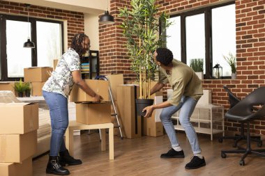 Heyecanlı çift yeni eve taşınıyor, ellerinde karton kutular ve mobilyasız oturma odasında saksı bitkileri var. Afrikalı Amerikalı erkek arkadaş ve kız arkadaş taşınırken yere paket koyuyorlar..