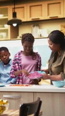 Dikey Afro-Amerikan ailesi evde yemek pişiriyor ve pizza gününün tadını çıkarıyor. Çocuklar ve ebeveynler taze malzemeler ekliyor ve kahkahaları paylaşırken lezzetli ev yapımı pizza yapıyorlar. Kamera A.