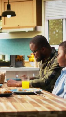 Vertical Video African American family serving breakfast together in a bright kitchen, parents pour orange juice and children enjoy eggs and toast. Cozy scene captures love and family bonding time