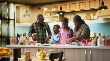 Neşeli Afrikalı Amerikalı ebeveynler çocuklara sebzeleri güvenli bir şekilde pişirmeyi ve kesmeyi öğretiyor, aileler eski tariflerden ev yapımı pizza yapmayı seviyor. Annem ve babam verimli yemek pişirme adımlarını anlatıyor. Kamera A.