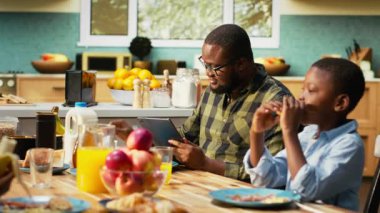 African american dad on tablet multitasking and chatting with his son, serving breakfast with his family and eating good homemade food in the kitchen. Togetherness and joy. Camera B.