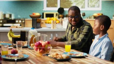 African American family serving breakfast together in a bright kitchen, parents pour orange juice and children enjoy eggs and toast. Cozy scene captures love and family bonding time. Camera B.