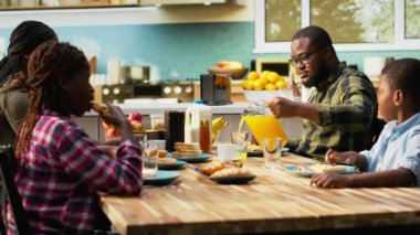 Black sweet family sharing croissants and orange juice in the morning, serving a rich colorful breakfast together under morning light. Young parents preparing the meal for their kids. Camera B.