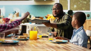 Happy family enjoying a homemade breakfast in the kitchen, serving omelettes, pastry and juice. Parents and children sharing laughter and the beauty of togetherness in a cozy morning. Camera B.