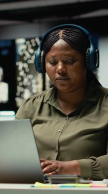 Dikey Videolu Kadın ofis çalışanı, gece odaklı kulaklıkla günlük işleri yönetirken, konsantrasyon ve verimliliğe yardımcı olacak ortam sesleriyle ilham buluyor. Kadın Çoklu Görev