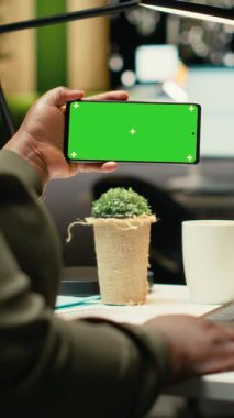 Masadaki akıllı telefondaki dikey video yeşil ekranda siyah iş adamı dizüstü bilgisayardaki performans ölçüm raporları üzerinde çalışıyor. Önemli verilerin toplanması ve gece yönetimsel görevlerin çözülmesi