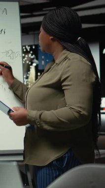 Dikey Video Afrikalı Amerikalı işçi, bilgi ve raporları özetliyor, veri analizi kullanarak şamdan çizelgeleriyle projeksiyonlar yapıyor. Gelir hedeflerine ulaşmak için öngörü. Kamera A