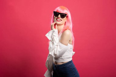 Siyah güneş gözlüklü ve elleri çenesinde poz veren neşeli genç bayan. Muhteşem mutlu kadın yüzüne dokunuyor ve modern stüdyoda pembe arka plana karşı güvenle duruyor..