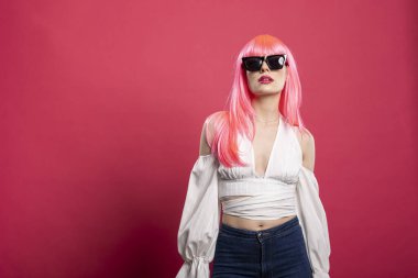 Siyah gözlüklü, beyaz elbiseli, güzel, neşeli bir kadın, pembe stüdyo geçmişine karşı güvenle gülüyor ve poz veriyor. Enerjik kadın neşe ve modern moda titreşimlerini ifade eder..