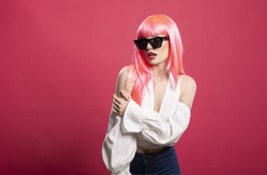 Modaya uygun güneş gözlüklü güzel genç bayan modern pembe bir stüdyoda poz veriyor. Beyaz bir bluz giyerek, bu şık portreye zarafet ve güven saçar..