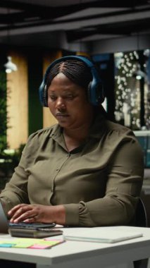 Dikey Videolu Kadın ofis çalışanı, gece odaklı kulaklıkla günlük işleri yönetirken, konsantrasyon ve verimliliğe yardımcı olacak ortam sesleriyle ilham buluyor. Kadın Çoklu Görev