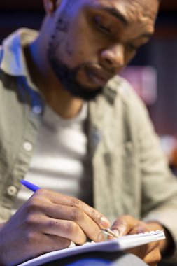 Erkek serbest çalışan, veri odaklı kayıtlar ve dosyalarla mali durumu gözden geçiriyor, evden çalışıyor ve ders kitabına not alıyor. Siyahi uzman bir geliştirme stratejisi üzerinde çalışıyor.