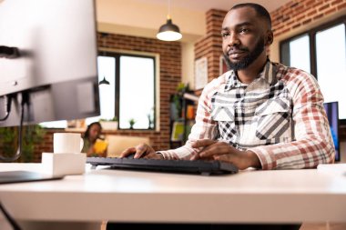 Evde çalışan kendine güvenen bir adam, şirket gelirlerini kontrol ediyor ve bilgisayardaki analizleri inceliyor. Erkek iş danışmanı modern tuğla duvar apartmanında proje sunumunu hazırlamak için masaüstü bilgisayarını kullanıyor.