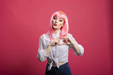 Pembe saçlı ve günlük kıyafetli güzel bir kadın göğsüne kalp jesti yapıyor, sıcaklık ve tutku saçıyor. Kadın model yalnız duruyor, şefkat ve şefkat paylaşıyor..