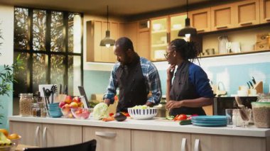 Afro-amerikan insanlar taze salata için bir kaseye doğranmış sebze ekliyorlar, kesme tahtasına organik sebzeler. Erkek arkadaş ve kız arkadaş vejetaryen yemeği yapıyorlar. Kamera A.