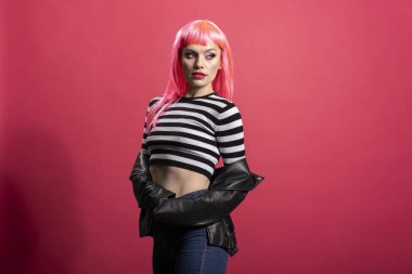 Parlak makyajlı ve renkli peruklu zarif bir bayan model çizgili üst ve deri ceket giyer, özgüvenle pembe arka planda modellik yapar. Stüdyoda poz veren alımlı şık kadının portresi..