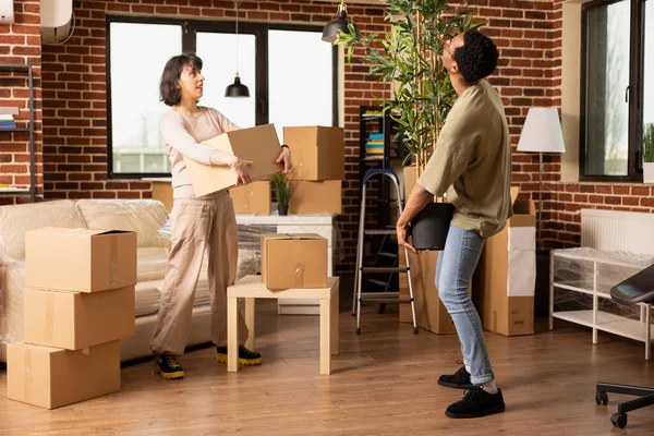 Kafkas kadın kahverengi kutuyu tutarken Afrikalı Amerikalı erkek arkadaşının saksı bitkisini yeni tuğla duvara taşımasını izliyor. Çok ırklı bir çift, eşyalarıyla birlikte taşınıyor..