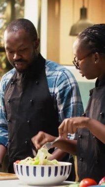 Dikey Video Siyah Adam aptalca davranıyor ve mimelerdeki şef gibi tuz ekliyor, salatayı kaseye koyup komik oluyor. Çift organik sebzelerle sağlıklı bir öğle yemeği hazırlıyor, dengeli bir yaşam tarzı.