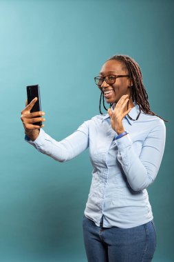 Gülümseyen siyahi kadın akıllı telefonuna el sallıyor, çevrimiçi izleyiciler için video kaydediyor ve dostane, samimi bir anı yakalıyor. Mutlu Afro-Amerikan bayan görüntülü arıyor, selamlama hareketi yapıyor..