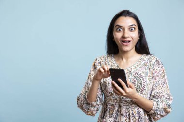 Sosyal medya uygulamaları ve web siteleri için akıllı telefonunu kullanan neşeli bir kadın, mavi arka planda sayfaları tarıyor. Genç bir kadın stüdyoda neşeli ve şaşkın, akıllı telefon..