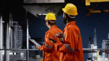 Turuncu ceketli ve kasketli bir erkek işçi, kıyı sondajındaki sistem operasyonlarını analiz ediyor, petrol çıkarma standartlarına uymayı ve platform güvenliğini sağlıyor. Kamera B.