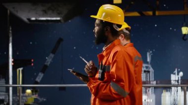 Turuncu ceketli ve kasketli bir erkek işçi, petrol ve gaz sanayii standartlarına uyarak kıyı sondajındaki sistem operasyonlarını analiz ediyor. Kamera B.