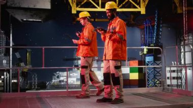 Turuncu ceketli ve kasketli bir erkek işçi, kıyı sondajındaki sistem operasyonlarını analiz ediyor, petrol çıkarma standartlarına uymayı ve platform güvenliğini sağlıyor. Kamera A.