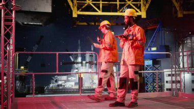 Turuncu ceketli ve kasketli bir erkek işçi, petrol ve gaz sanayii standartlarına uyarak kıyı sondajındaki sistem operasyonlarını analiz ediyor. Kamera A.