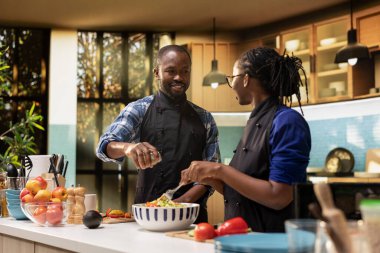 Siyah erkek arkadaş kavanozdan salataya baharat ve sos ekliyor. Organik öğle yemeğini taze biyolojik malzemelerle tatlandırıyor. Kız arkadaşın sebzeleri bir kasede karıştırıyor, vejetaryen yemeği..