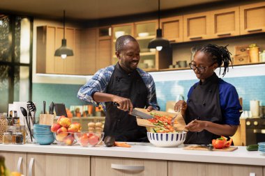 Siyah çift taze salata için bir kaseye doğranmış sebze ekliyor, kesme tahtasında birden fazla organik sebze var. Erkek arkadaş ve kız arkadaş vejetaryen yemeği yapıyor, yaşam tarzı....