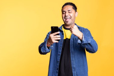 Hintli adam sakin ve gülümsüyor, akıllı telefonuyla etkileşimde, dijital uygulamalarda geziniyor ve kablosuz bağlantının keyfini çıkarıyor. Parlak turuncu bir stüdyonun arka planında sıradan bir poz.