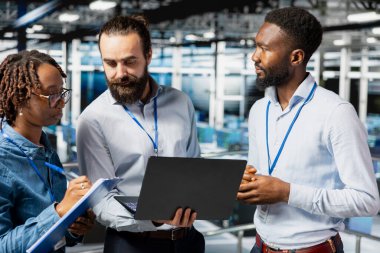 Data center team leader and workers reviewing performance dashboards of AI equipment rigs. Server room engineers overseeing machine learning algorithms on laptop, elevated workstation.