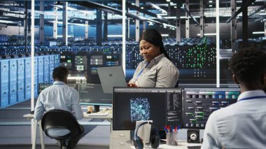 Düz ağaç programlaması yapmak için dizüstü bilgisayar kullanan veri merkezi yönetimi, kaynak tahsisini optimize ediyor. Sunucu odasındaki kadın, bakım planlama için öngörücü yapay zeka görselleştirme sistemleri kullanıyor, kamera A