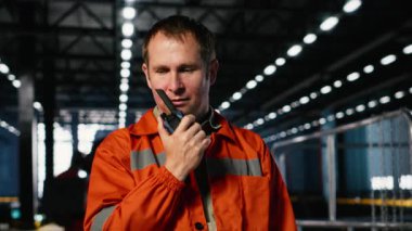 Endüstri işçisi personel ile iletişim kurmak için telsiz kullanıyor, depoda ağır makinelerin yanında duruyor. Sanayi fabrikasında üretim sürecini destekleyen işgücü. Kamera B.