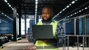 Bir depo tesisinde metal aletlerle çevrili Afrikalı Amerikalı teknisyen dizüstü bilgisayarla çalışıyor ve profesyonel işçileri gösteriyor. Ürün yaratmak için güç üretiyorum. Kamera B.